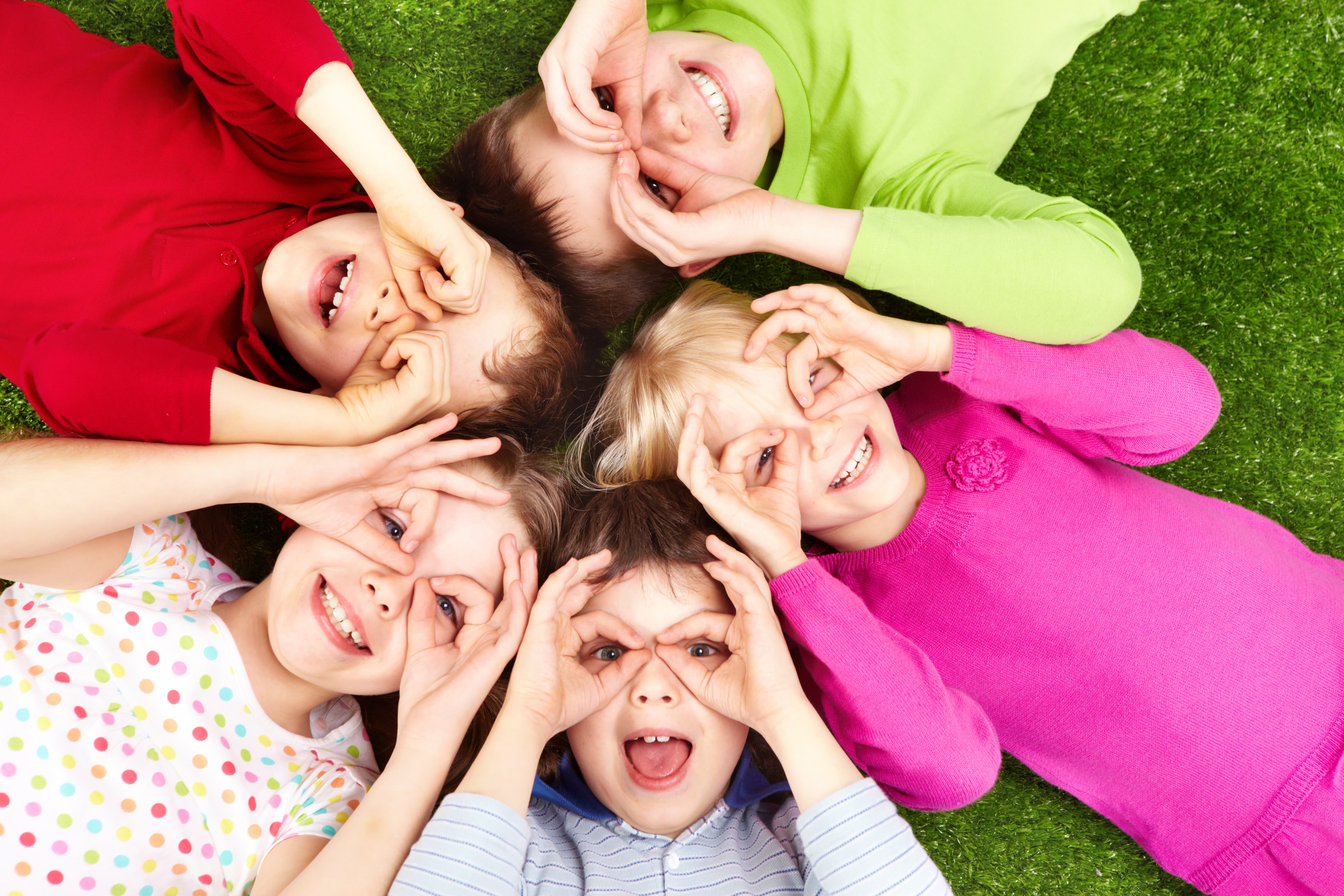 A group of children laying in the grass while making fake glasses on their eyes with their hands.